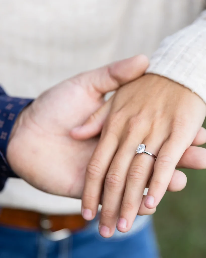 Close-up of a couple's hands held together, showing a classic solitaire oval diamond engagement ring on a silver band.