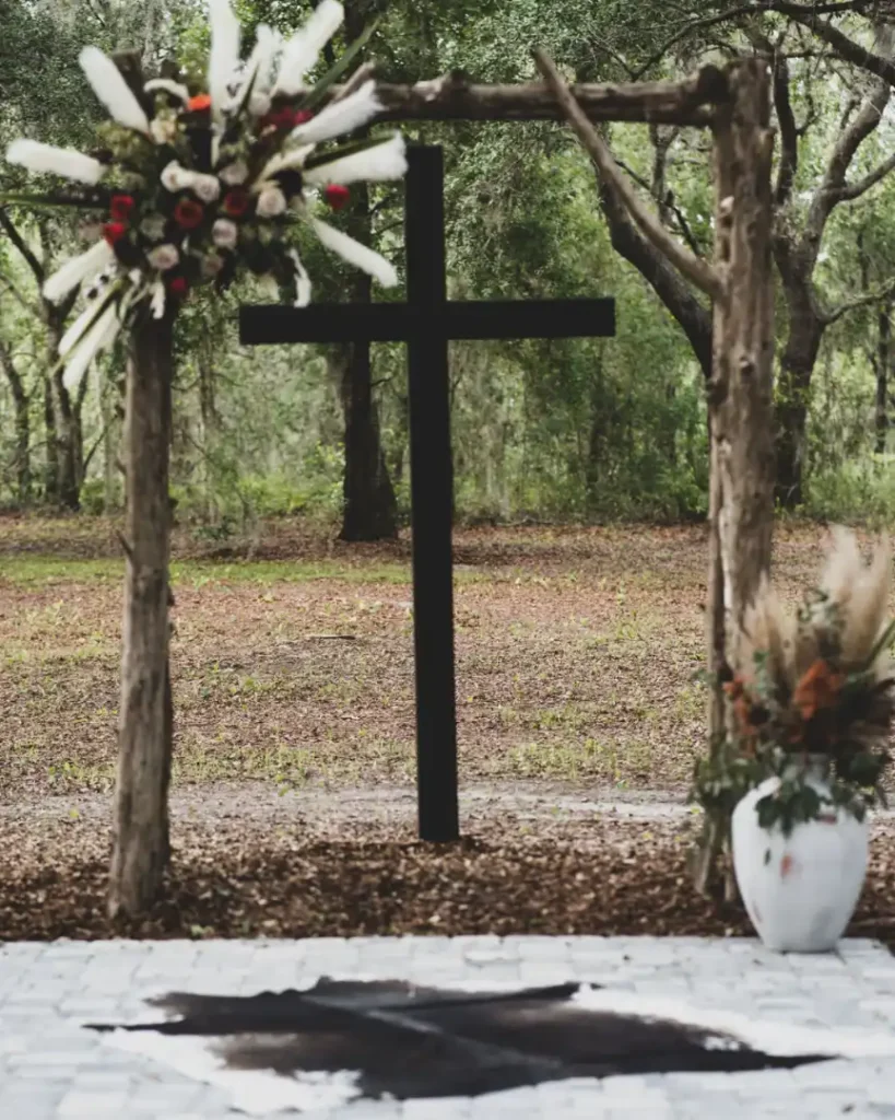 Outdoor rustic wedding arch decorated with white floral arrangements and a central dark wooden cross.