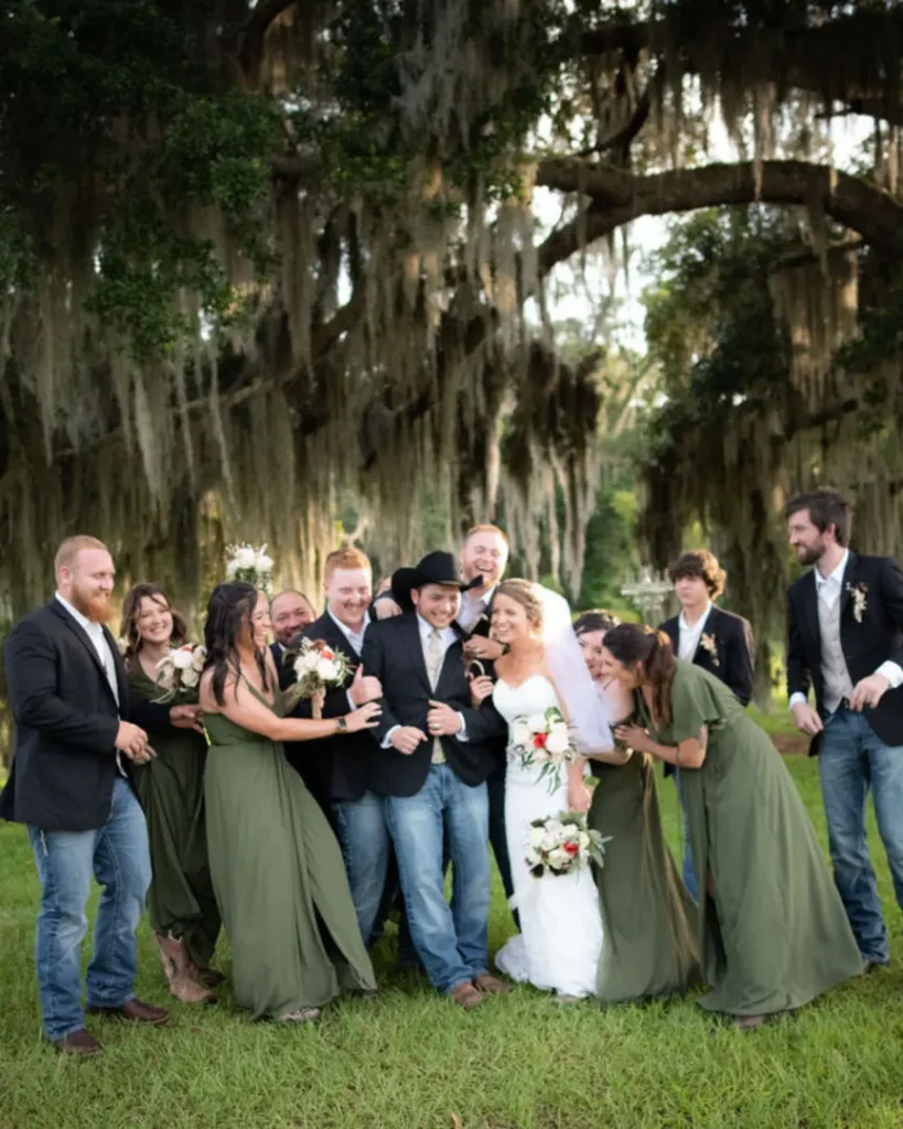A bride and groom laughing and celebrating outdoors with their bridal party, surrounded by moss-draped trees.
