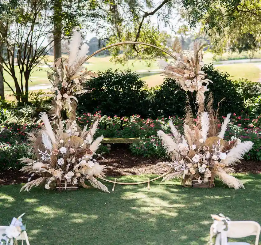 Large wooden circular wedding arch adorned with pampas grass, white flowers, and lush greenery in an outdoor garden setting.