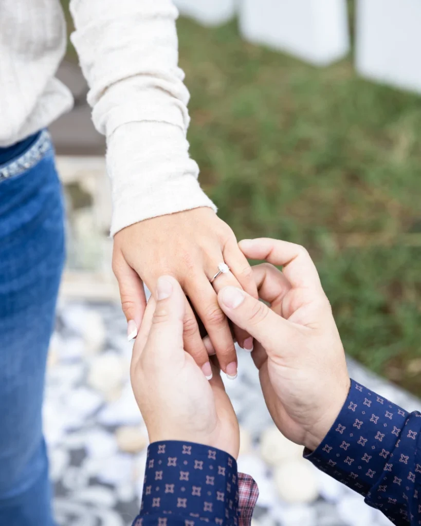 A close-up of a man placing an engagement ring on a woman's finger as they hold hands.
