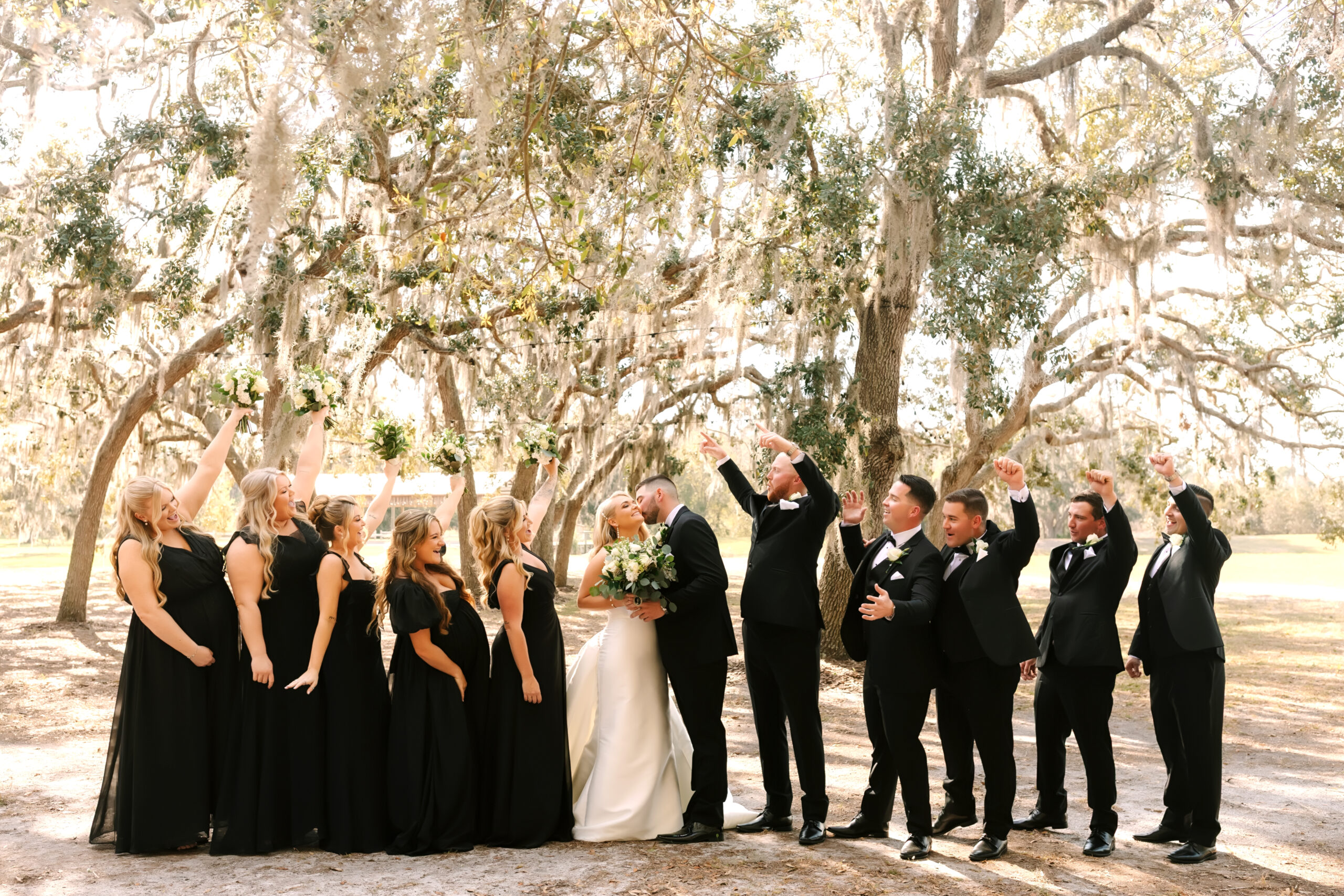 Bridal party in black attire posing together under oak trees draped in Spanish moss