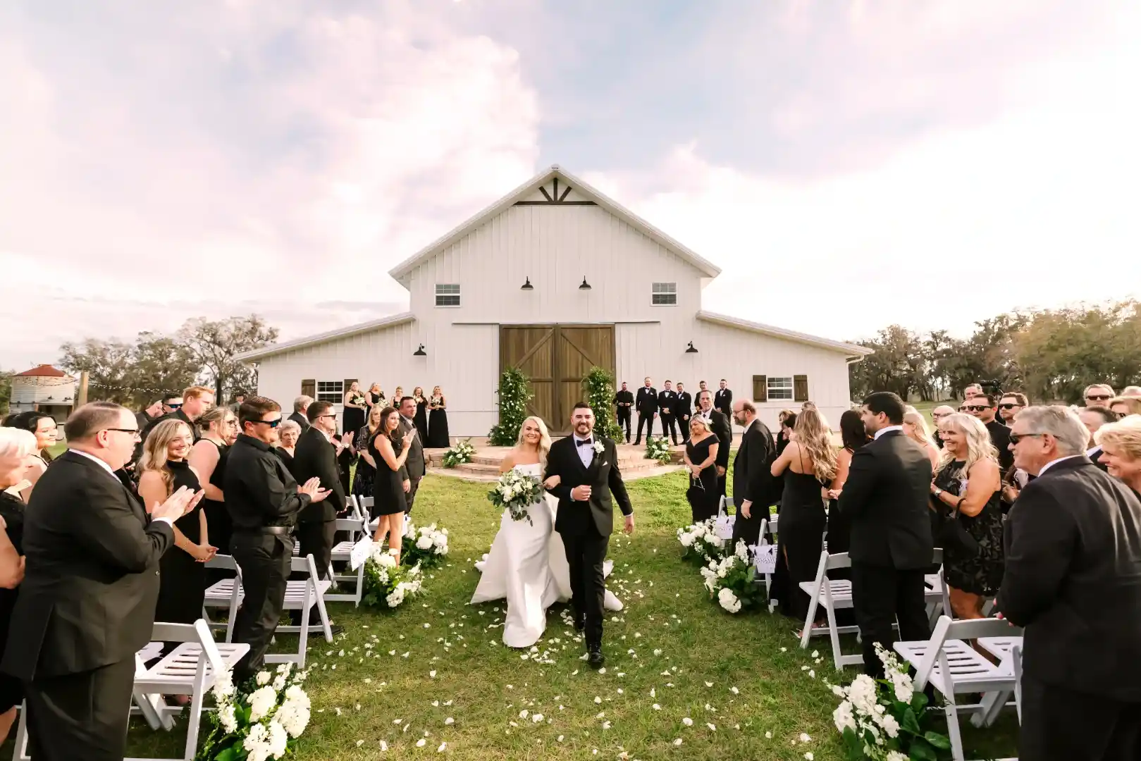 Bride and groom walking down the outdoor aisle smiling as seated guests in black attire applaud in front of a white wedding barn