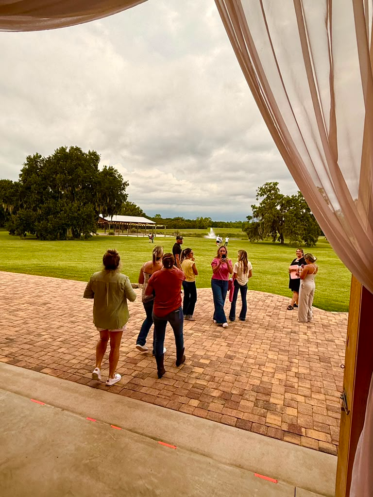 Guests gather on a grassy lawn with trees and a draped outdoor structure at an open-air wedding venue.