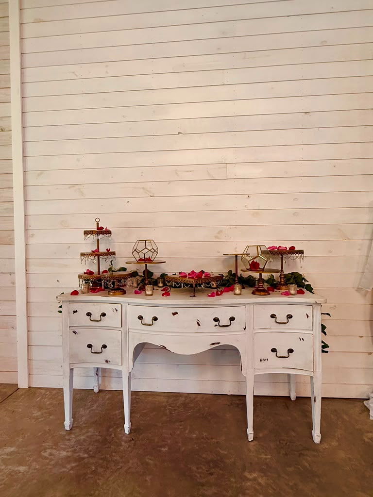 Vintage white dresser with floral decor and photo frames set up as a guest book table against a wood wall.